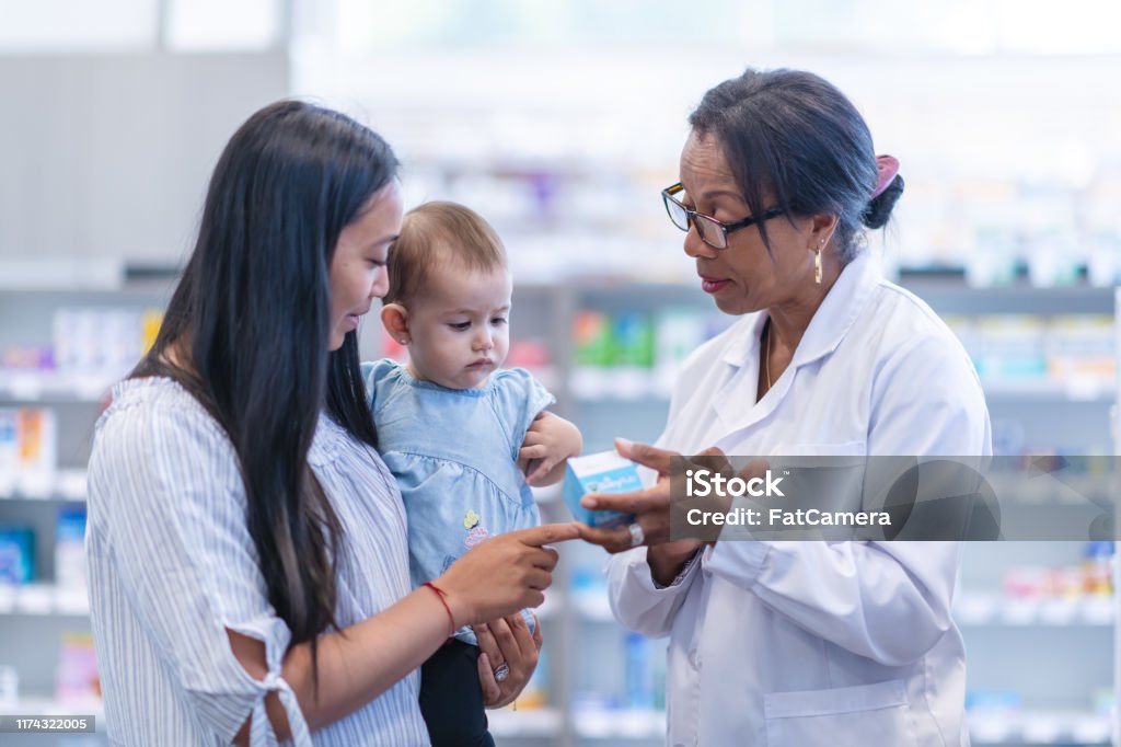 A young mother of Asian descent is picking up a prescription. The woman is holding her baby girl while at the store. She is speaking to a female pharmacist of African descent about the medicine